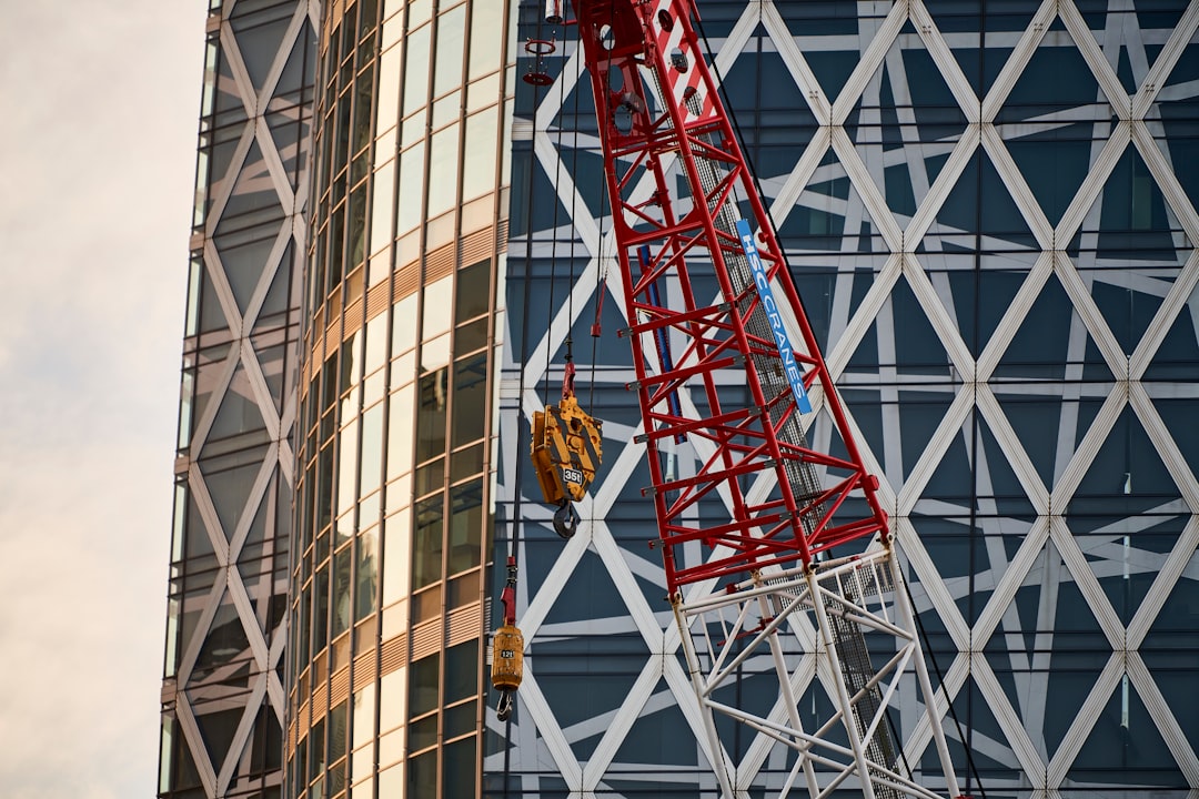 A huge construction crane rests against the glass wall of a modern skyscraper nearing completion. The cool, geometrically patterned façade of the building contrasts sharply with the cranes rugged steel frame, painted a vibrant red. This is the dynamic moment when a city is growing and the landscape of the future is taking shape. This photograph is filled with the creative and developmental energy inherent in the act of construction.